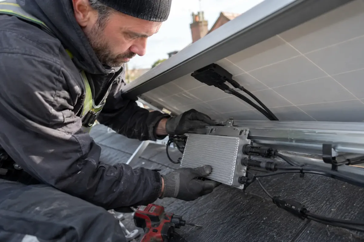 Installer replacing a small finned metal microinverter on a mounting rail beneath a solar panel.